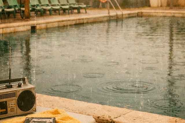 Piscina municipal vacía bajo la lluvia en Mallorca, con altavoces oxidados y círculos en el agua, evocando la melancolía de la canción Ciutat de Jaume Escala.