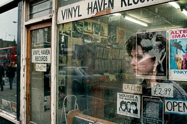 Reflejo borroso de una joven en el cristal sucio de una tienda de discos de los 80, con interior visible detrás y estética cruda de carrete